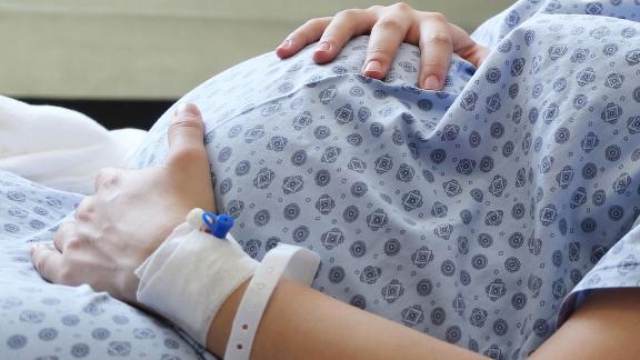 A close up of a pregnant woman in a hospital gown, clutching her stomach.