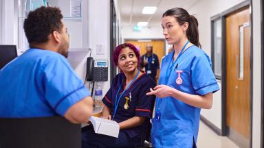 Healthcare staff confer at a nurses’ station in a hospital corridor, with notes and equipment nearby.
