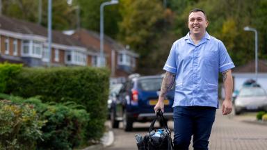 A healthcare worker in uniform walks along a residential street carrying a work bag.