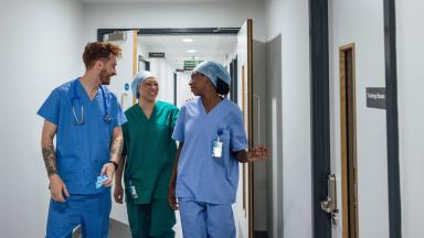 Three healthcare workers in scrubs walking together along a hospital corridor, talking as they pass open rooms.
