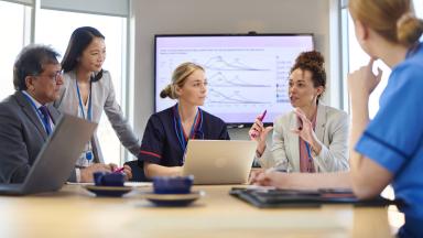 Group of healthcare professionals seated around a table discussing information, with a screen displaying charts in the background.