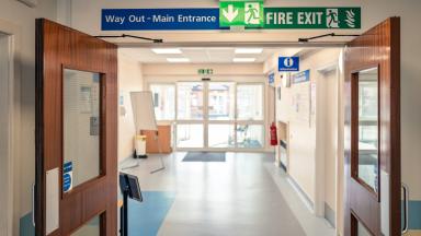 Hospital corridor with double doors and overhead signs reading “Way Out – Main Entrance” and “Fire Exit.”