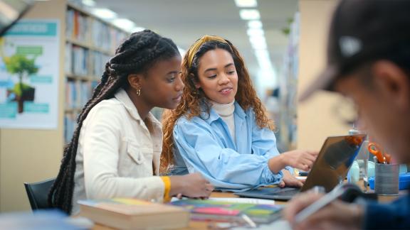 Two students studying together on a laptop in a library with books and notes