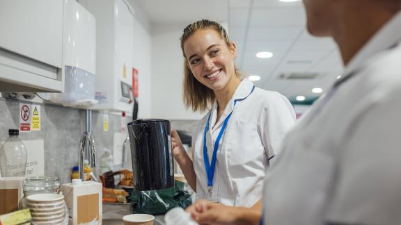 Healthcare worker holding a kettle in a staff kitchen with cups, containers and safety notices on the wall