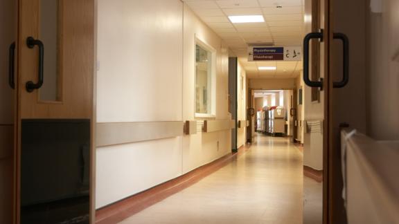 Long hospital corridor with open doors, overhead signage for physiotherapy, and equipment trolleys visible at the far end