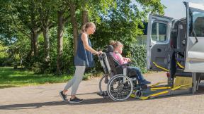 A carer pushing a person in a wheelchair up a ramp and into a car.