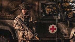 Armed forces personel stood in front of a land rover ambulance with their rifle at a stand gaurd position.
