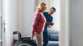 A care worker supports an older person as they stand beside a wheelchair in a bright room.