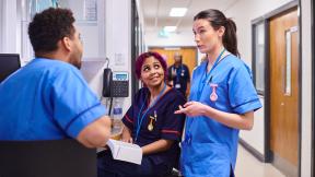 Healthcare staff confer at a nurses’ station in a hospital corridor, with notes and equipment nearby.