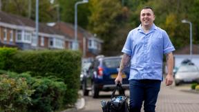 A healthcare worker in uniform walks along a residential street carrying a work bag.