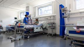 A nurse speaks with a patient lying in a hospital bed on a bright ward with medical equipment and monitoring devices.