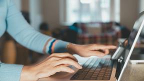 Close-up of hands typing on a laptop keyboard at a desk in an indoor setting.