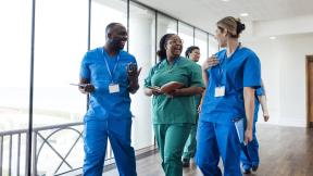 Group of healthcare workers in scrubs walking together along an indoor corridor while talking and holding notebooks.