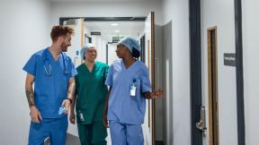 Three healthcare workers in scrubs walking together along a hospital corridor, talking as they pass open rooms.