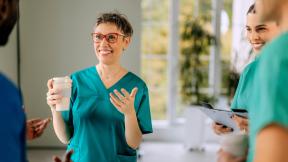 Healthcare workers in scrubs standing together indoors, talking while one holds a takeaway cup.
