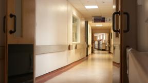 Long hospital corridor with open doors, overhead signage for physiotherapy, and equipment trolleys visible at the far end