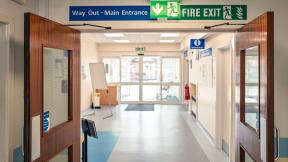 Hospital corridor with double doors and overhead signs reading “Way Out – Main Entrance” and “Fire Exit.”