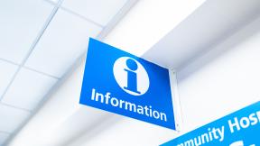 Blue overhead hospital sign with a white information symbol and the word “Information,” mounted on a ceiling bracket in a bright corridor.