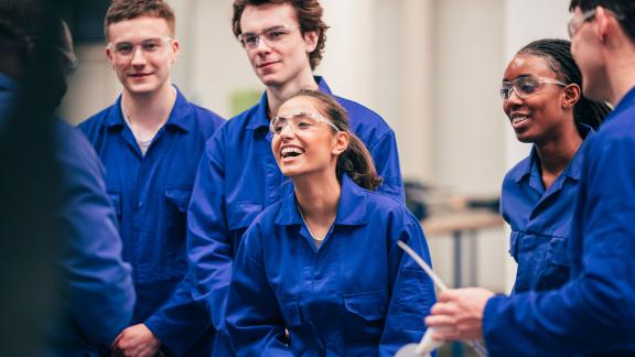 A diverse group of apprentices, wearing boiler suits, being taught.