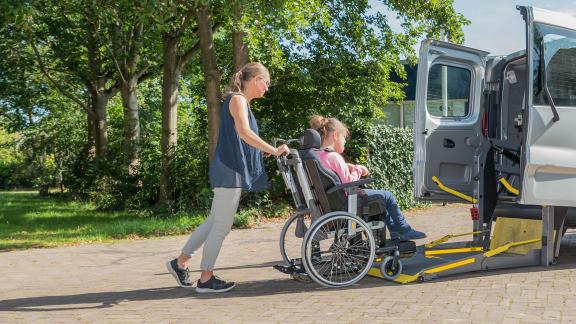 A carer pushing a person in a wheelchair up a ramp and into a car.