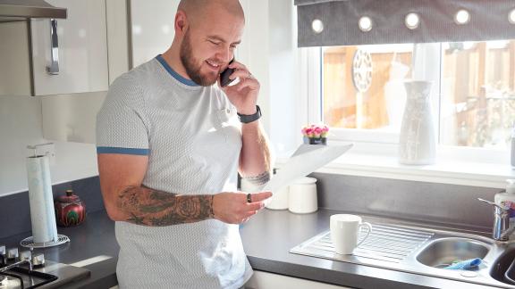 A smiling man in a kitchen, making a phone call. 