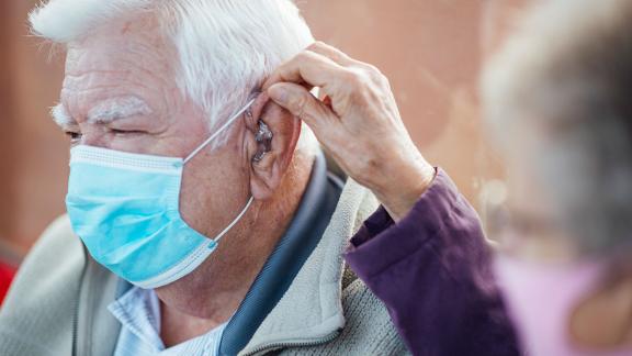 A masked patient having his hearing aid adjusted by his wife.