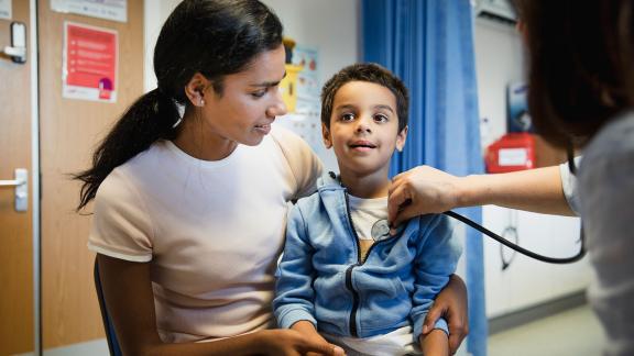 A boy sitting on his mother's knee while the doctor checks his heart rate.