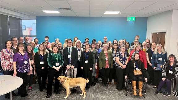 Large diverse group of delegates posing for a picture in a office building room. Among the group, Matthew James standing with a blonde Labrador. 