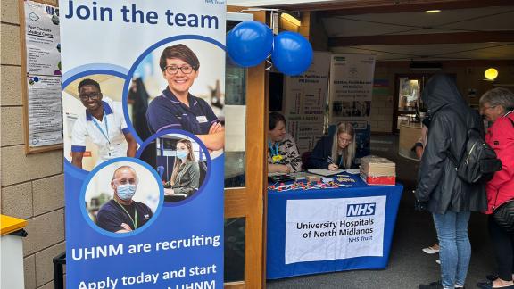 People at a table with a sign saying "Join the Team". A group of individuals gathered around a table, speaking to the members of staff at University Hospitals of North Midlands..