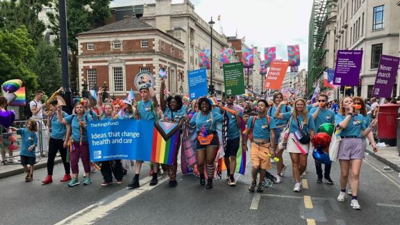 A diverse group of individuals walking down a closed street in London, holding vibrant rainbow flags and a banner with "The Kings Fund: Ideas that change health and care". .