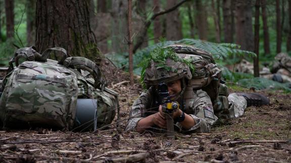 Armed forces personel in a prone position in a wooded area pointing their rifle towards the camera with their gear off to one side.