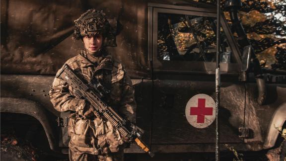 Armed forces personel stood in front of a land rover ambulance with their rifle at a stand gaurd position.