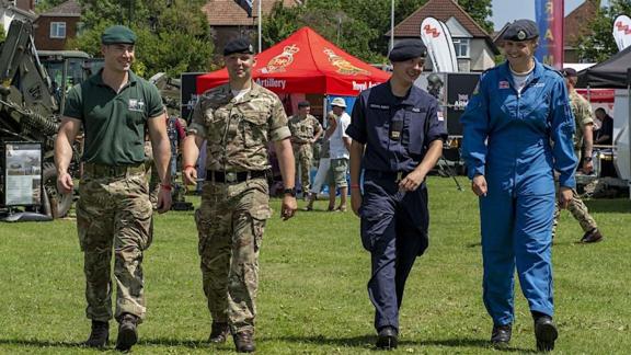 A group of armed forces personel walking through an event during summer. There are 4 indirviduals in uniform representing the Royal Marines, Army, Navy and Air Force.