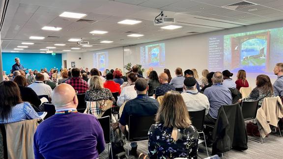 A large group seated in an auditorium facing two projector screens with a speaker talking them through the slides.