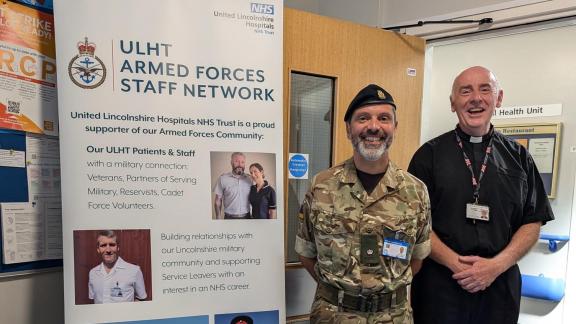 Two people are posing for an image in front of a pull up banner wearing army personnel uniform. The banner contains ULHT Armed Forces Staff Network information on.
