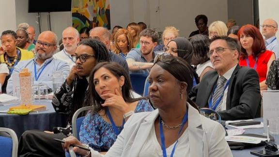 A diverse group of delegates, representing various backgrounds, attentively listens to a speaker at a conference. 