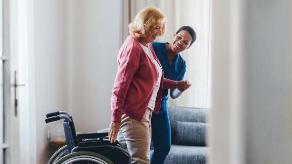 A care worker supports an older person as they stand beside a wheelchair in a bright room.