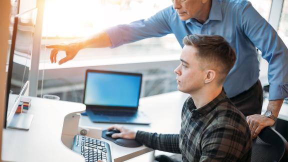 A supervisor stands beside a colleague at a desk, pointing to information on a desktop computer
