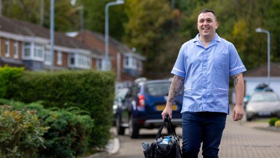 A healthcare worker in uniform walks along a residential street carrying a work bag.