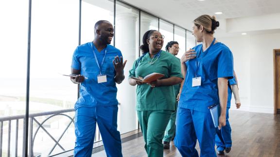 Group of healthcare workers in scrubs walking together along an indoor corridor while talking and holding notebooks.