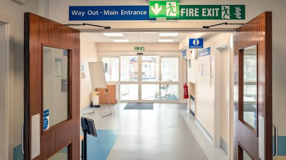 Hospital corridor with double doors and overhead signs reading “Way Out – Main Entrance” and “Fire Exit.”