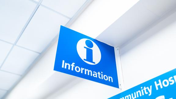 Blue overhead hospital sign with a white information symbol and the word “Information,” mounted on a ceiling bracket in a bright corridor.