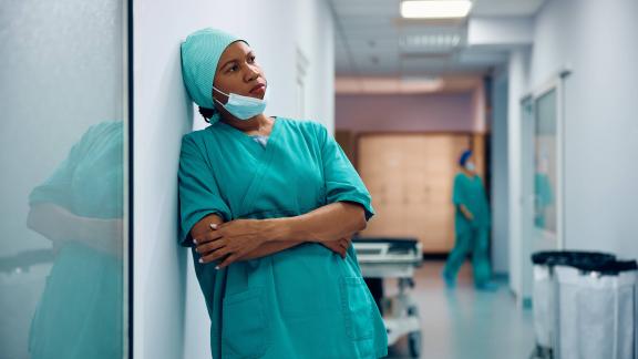 Medical staff in green scrubs leaning against a wall in a hospital corridor, with another staff member and equipment trolley in the background.