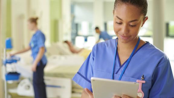 Nurse in a ward area using a tablet device, with hospital beds and staff visible in the background.