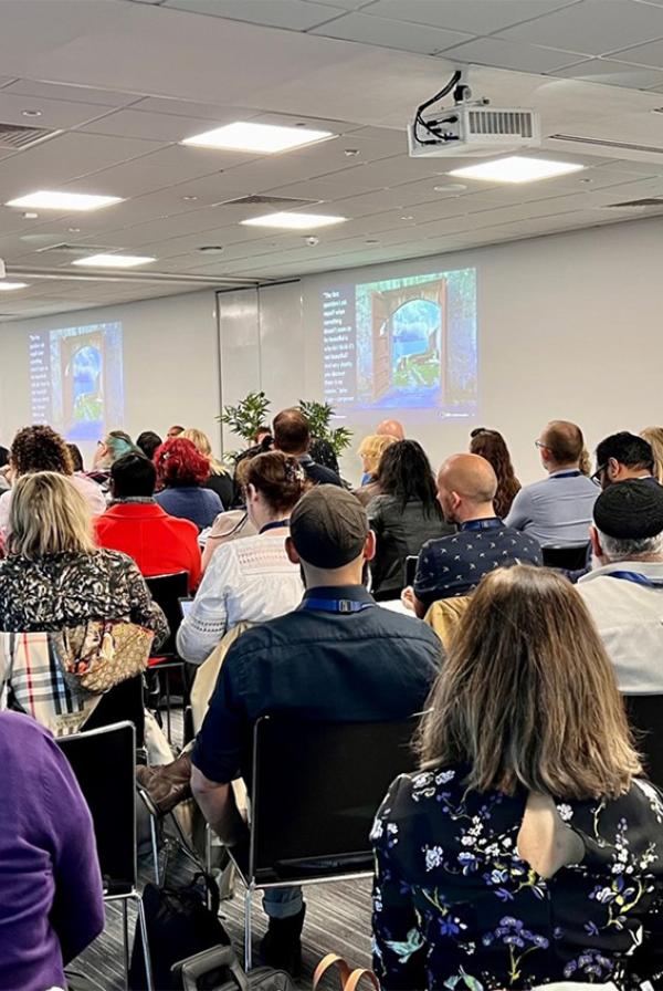 A large group seated in an auditorium facing two projector screens with a speaker talking them through the slides.