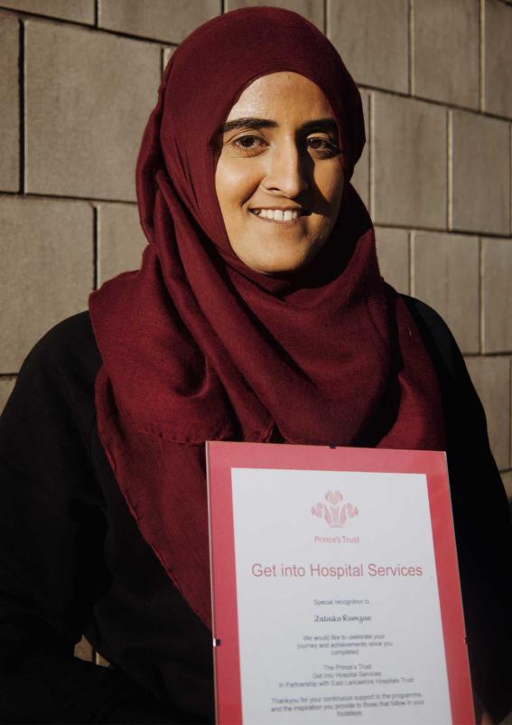 A young individual posing for a photograph with their certificates from the Princes' Trust in front of a brink background.